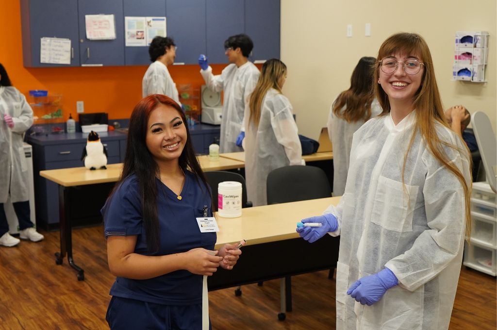 Phlebotomy students in a classroom setting, practicing skills and smiling in lab coats and gloves.